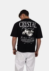 Zwarte katoenen t-shirt met in het groot witte tekst "CRYSTAL Paris" op de achterkant, vergezeld van diamantillustraties en wasinstructies eronder.