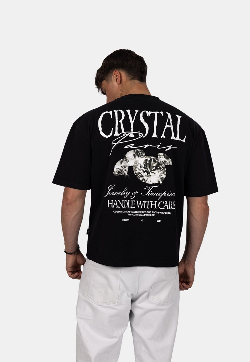 Zwarte katoenen t-shirt met in het groot witte tekst "CRYSTAL Paris" op de achterkant, vergezeld van diamantillustraties en wasinstructies eronder.