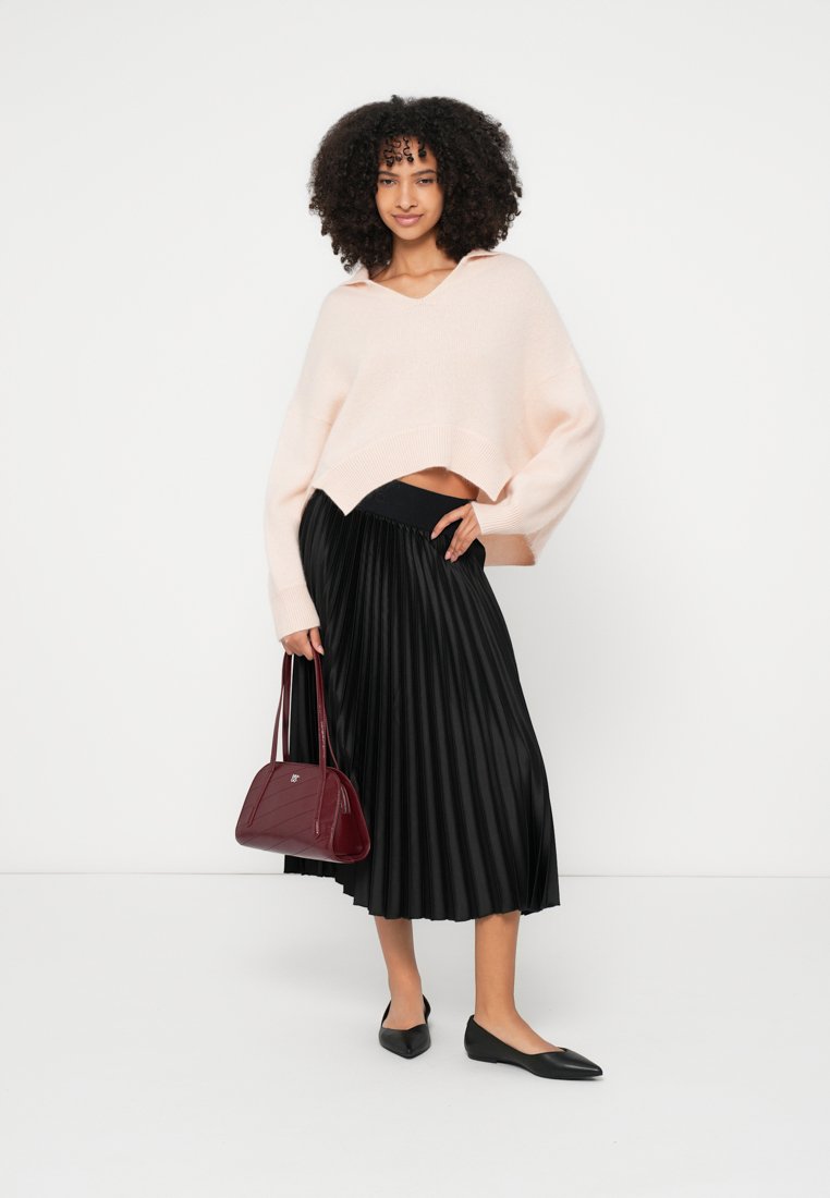 Pull court en tricot rose clair, jupe midi noire plissée, ballerines noires et sac à main bordeaux au design structuré.