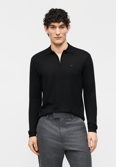 Homme aux cheveux bouclés portant une chemise noire à manches longues avec fermeture éclair et un pantalon de costume gris, sur un fond blanc uni.