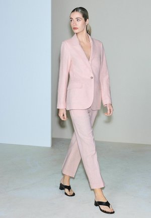 Femeie care merge purtând un costum roz deschis croitorit, cu pantofi negri cu vârf decupat, pe un fundal simplu gri și alb.