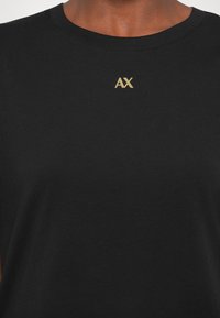 Čierne bavlnené tričko s okrúhlym výstrihom. Na hrudi sa nachádza malé zlaté logo „AX“. Mäkká textúra, jednoduchý dizajn.