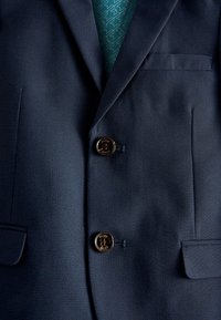Dark blue textured blazer with two brown buttons labeled "Baker," featuring flap pockets and a patterned teal inner lining visible.