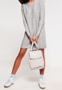 Ljusgrå ryggsäckshandväska med en strukturerad design, slät textur, klaffstängning och korta handtag. Bärs med en grå tröjdress och vita sneakers.