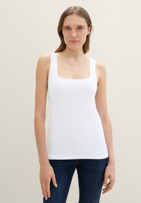 White fitted tank top with a square neckline and wide shoulder straps, made from smooth fabric. Paired with dark blue jeans.