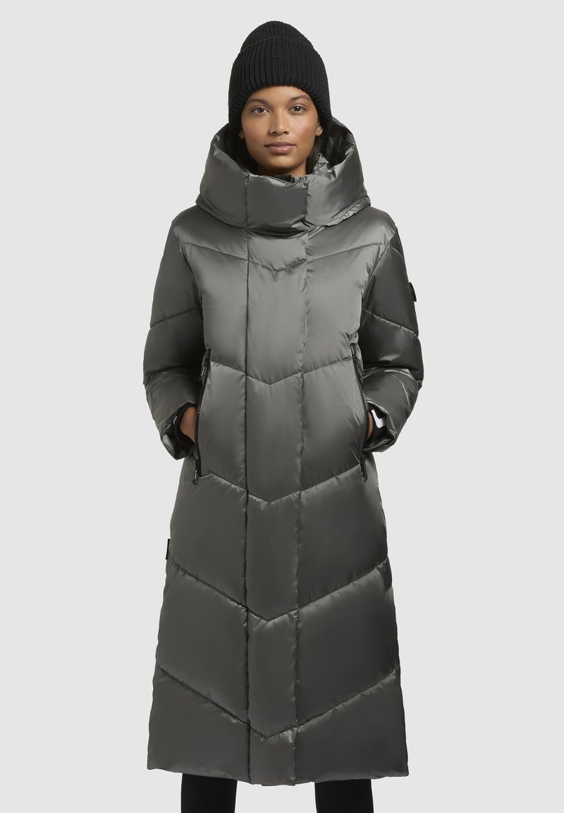 Lange, grijze pufferjack met chevronpatroon, hoge kraag en zakken. Model draagt een zwarte gebreide beanie. Strak, glanzend materiaal.