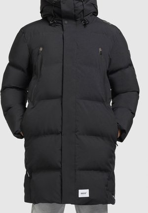 Manteau d'hiver matelassé noir avec capuche, poches zippées avant et mains dans les poches latérales, porté sur un pantalon gris clair.