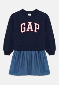 Tengerészkék pulóver piros és fehér "GAP" logóval, világoskék farmer szoknyával kombinálva. Rugalmas mandzsettái és rakott szoknya dizájnja van.