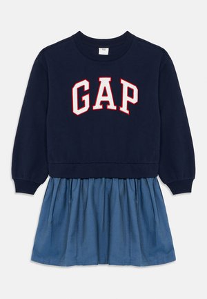 Felpa navy con logo "GAP" rosso e bianco, abbinata a una gonna di jeans leggera blu chiaro. Presenta polsini a coste e un design di gonna arricciata.