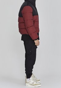 Rød og sort dunjakke med quiltede paneler, lynlåslukning og elastiske manchetter, parret med sorte cargobukser og beige sportssko.