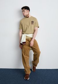 Beige katoenen t-shirt met een zwarte patch, gecombineerd met bruine corduroy broek. Witte sweater rond de taille geknoopt en bruine sneakers.