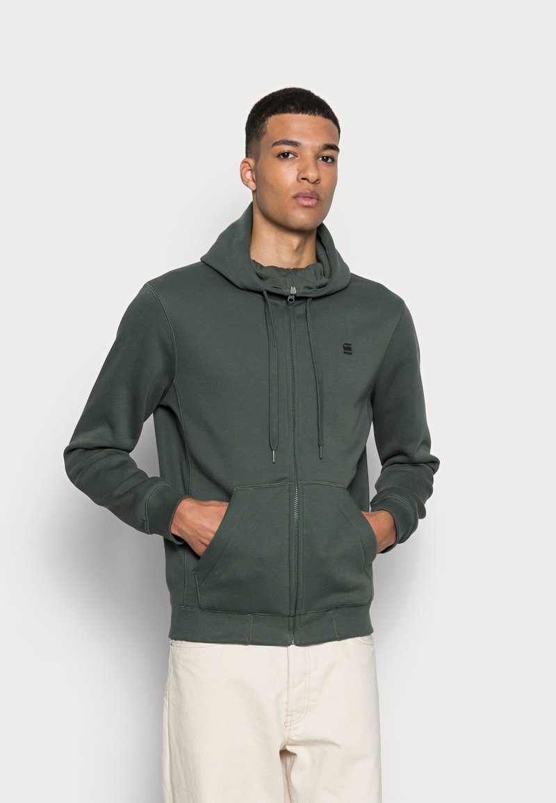 Veste à capuche zippée verte avec un cordon, poches avant, poignets côtelés et un accent logo sur la poitrine. Fabriquée en tissu doux et texturé.