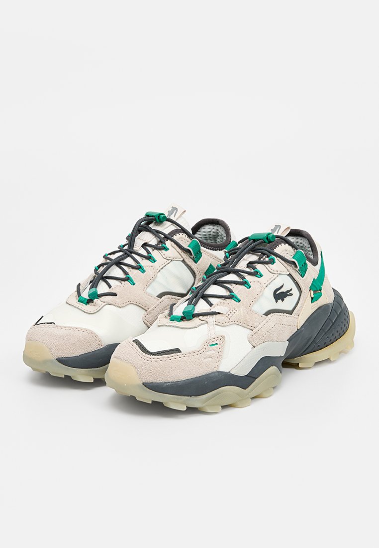 Sneakers Lacoste w połączeniu beżowego zamszu i białego materiału syntetycznego, z zielonymi akcentami, czarnymi sznurowadłami i teksturowaną gumową podeszwą.
