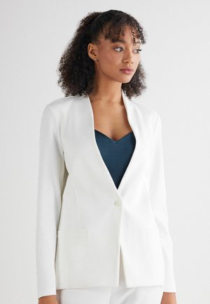 Weißer Blazer mit strukturiertem Design, Ein-Knopf-Verschluss und zwei Vorderaschen. Getragen über einem dunkelblauen V-Ausschnitt-Oberteil. Glatte Textur.