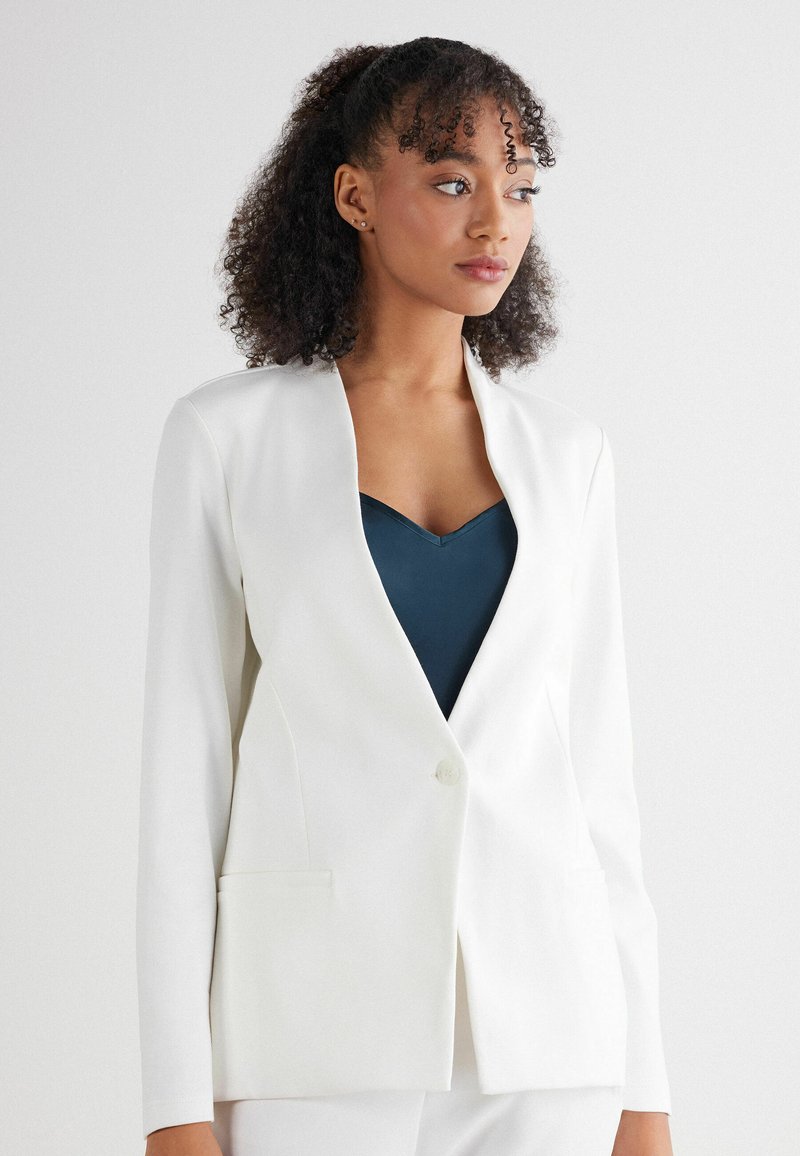 Witte blazer met een gestructureerd ontwerp, enkele knoopsluiting en twee voorkeurszakken. Draag over een donkerblauw V-hals shirt. Gladde textuur.