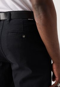 Pantaloni negri din material, cu o buzunar posterior cu nasture. O curea neagră din piele este purtată la talie, vizibilă în apropierea vârfului.