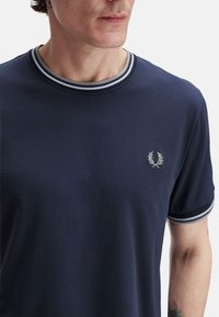Camiseta azul marino de algodón, con un cuello redondo acanalado con rayas en gris claro y blanco, y un pequeño logo bordado en el pecho.