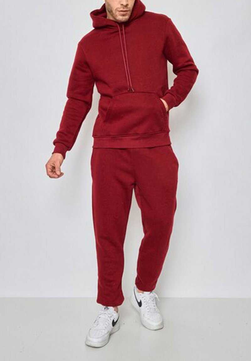 Un pulled rouge avec une poche avant et des cordons ajustables, associé à un pantalon de jogging rouge assorti. Porté avec des baskets blanches.