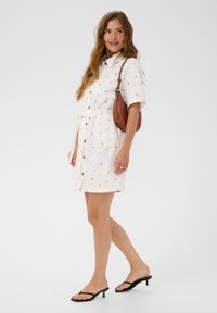 Femme portant une robe blanche à manches courtes avec des boutons et un petit imprimé floral, portant un sac à bandoulière marron et des sandales à talons noires, marchant.