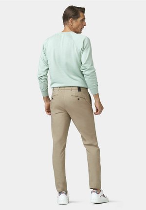 Mann trägt hellblauen Pullover, beige Hose und weiße Sneakers, steht leicht seitlich gedreht vor einfarbigem Hintergrund.