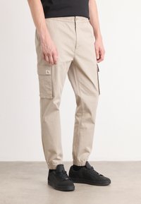 Beige cargobroek met een elastische tailleband, zijzakken en ribgebreide boorden, gecombineerd met zwarte sneakers op een neutrale achtergrond.