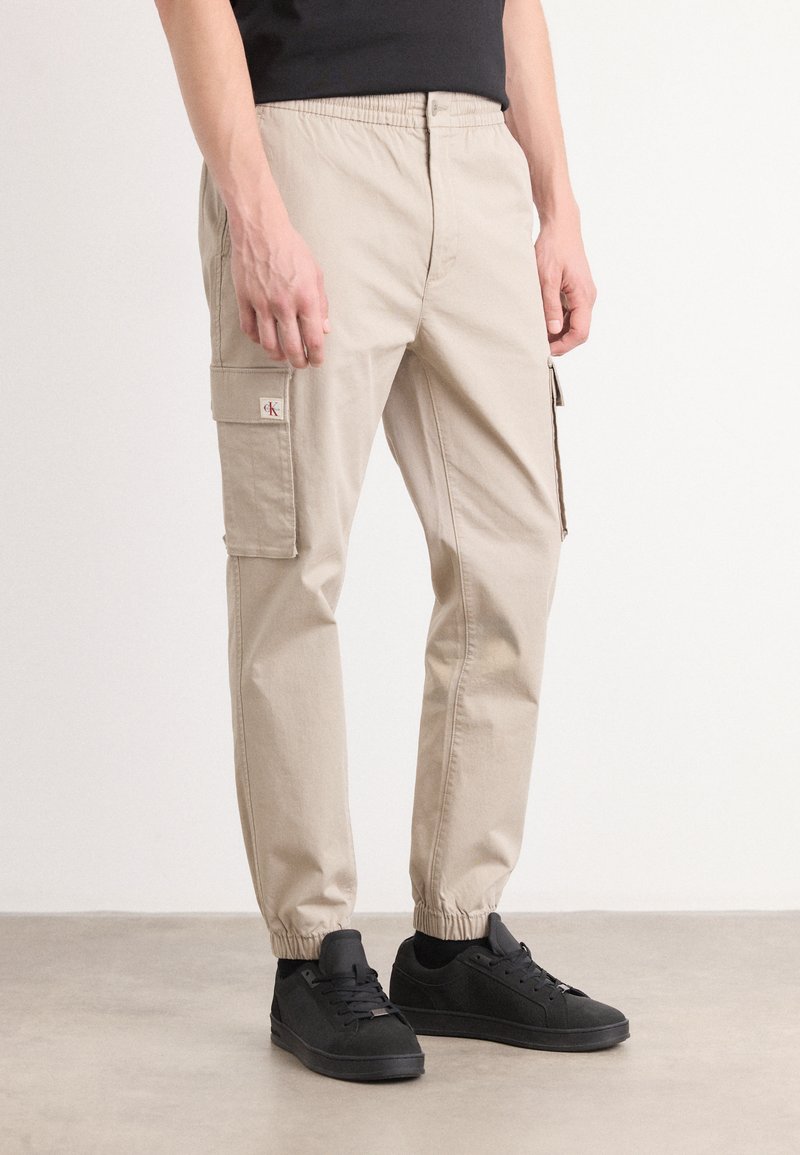 Beige cargobroek met een elastische tailleband, zijzakken en ribgebreide boorden, gecombineerd met zwarte sneakers op een neutrale achtergrond.