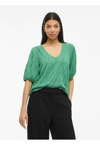 Top verde, strutturato, con scollo a V e maniche corte a sbuffo. Abbinato a pantaloni neri su misura. Tessuto liscio con un design a pieghe.