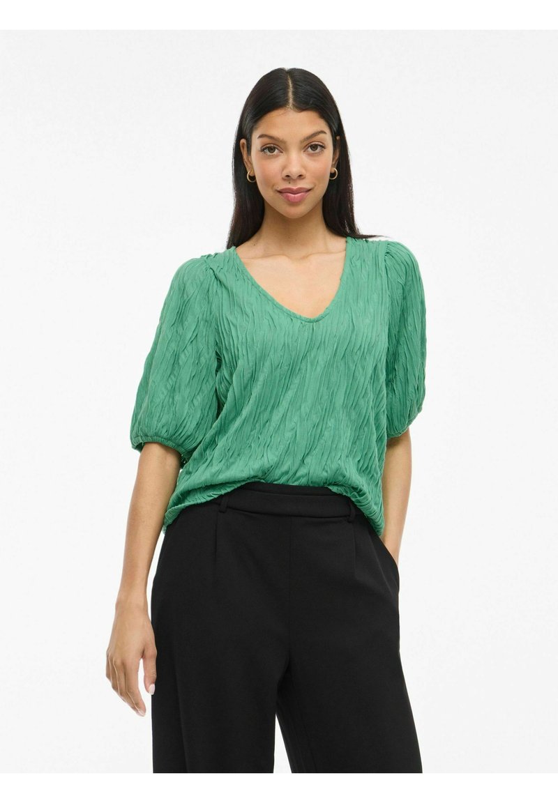 Top verde, strutturato, con scollo a V e maniche corte a sbuffo. Abbinato a pantaloni neri su misura. Tessuto liscio con un design a pieghe.