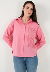Pink long-sleeve shirt with a striped texture, featuring a lace pocket with fringe detail. Button-up design and classic collar.