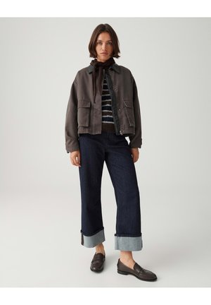 Femme portant un jean large court foncé avec de grands revers, un pull rayé, une veste sombre, une écharpe marron et des chaussures marron sans lacets.