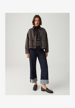 Frau trägt dunkle, hochgekrempelte, weite Jeans mit großen Manschetten, einen gestreiften Pullover, eine dunkle Jacke, einen braunen Schal und braune Slipper.
