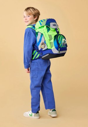 Young child in blue jumpsuit smiling, wearing a large green and blue striped backpack with safety reflectors, standing on beige background.