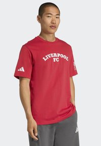 Muž oblečený v červenom tričku Liverpool FC s bielym textom a logom, doplnený o sivé tepláky Adidas na jednoduchom svetlom pozadí.