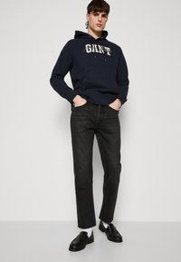 Felpa con cappuccio blu navy con logo bianco, abbinata a jeans scuri e mocassini neri. Il modello è in piedi con le mani in tasca su uno sfondo semplice.