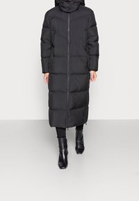 Svart, pösig lång kappa med huva och framdragkedja. Har chevron-quilting och elastiska ärmslut. Bärs över svarta leggings och stövlar.