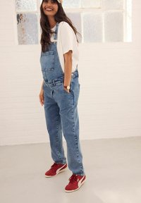 Pantalones de mezclilla en azul claro con tirantes ajustables y un bolsillo frontal, combinados con una camiseta blanca y zapatillas rojas. Textura suave.