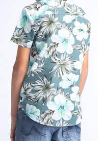 Camicetta a maniche corte, con bottoni, di colore teal con un motivo floreale tropicale caratterizzato da grandi fiori bianchi e blu chiaro e foglie verdi.