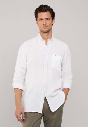 Weißes Leinen-Hemd mit Knöpfen und aufgekrempelten Ärmeln, ausgestattet mit einer Brusttasche und subtiler Textur. Kombiniert mit beigen Hosen für ein leichtes Outfit.