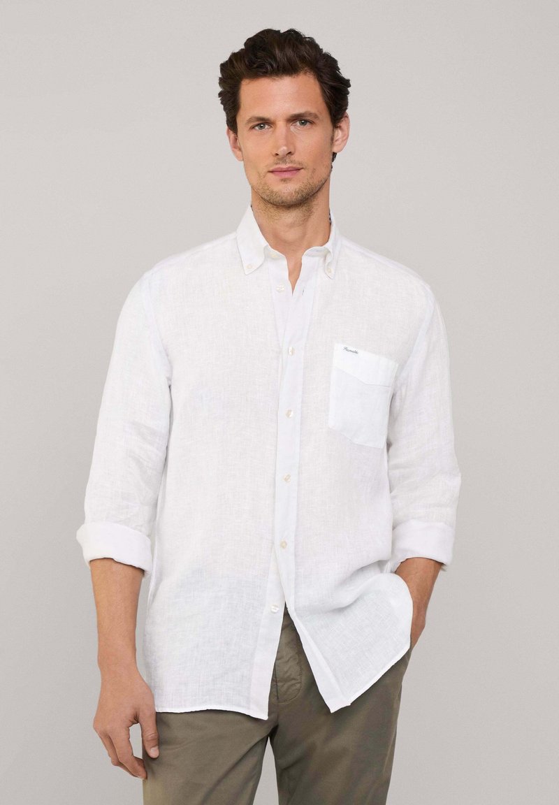 Camisa blanca de lino con botones y mangas arremangadas, que cuenta con un bolsillo en el pecho y una textura sutil. Combinada con pantalones beige para un atuendo ligero.