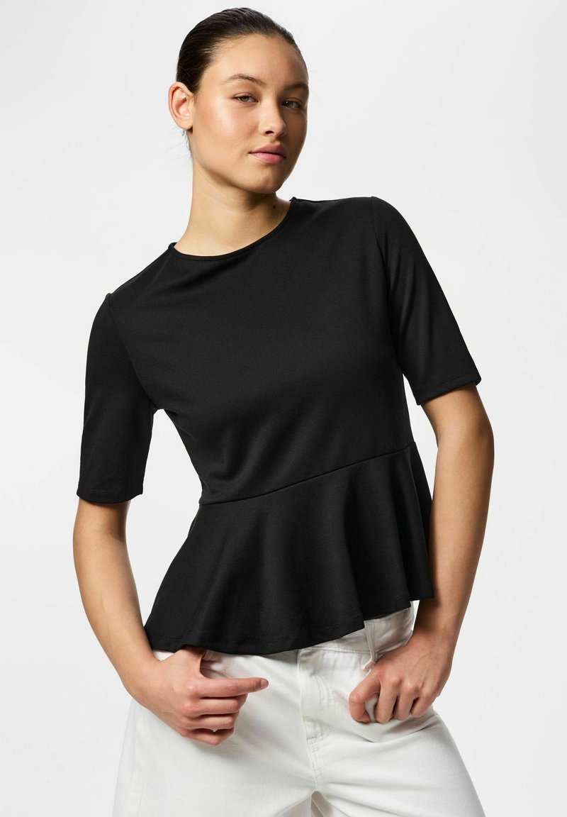 Jeune femme portant un top peplum noir à manches courtes et un pantalon blanc, debout avec les mains près de la taille sur un fond uni.