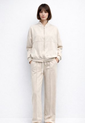 Ung kvinde stående med hænderne i lommerne iført beige lynlåstrøje og matchende bukser med vide ben foran en ensfarvet hvid baggrund.