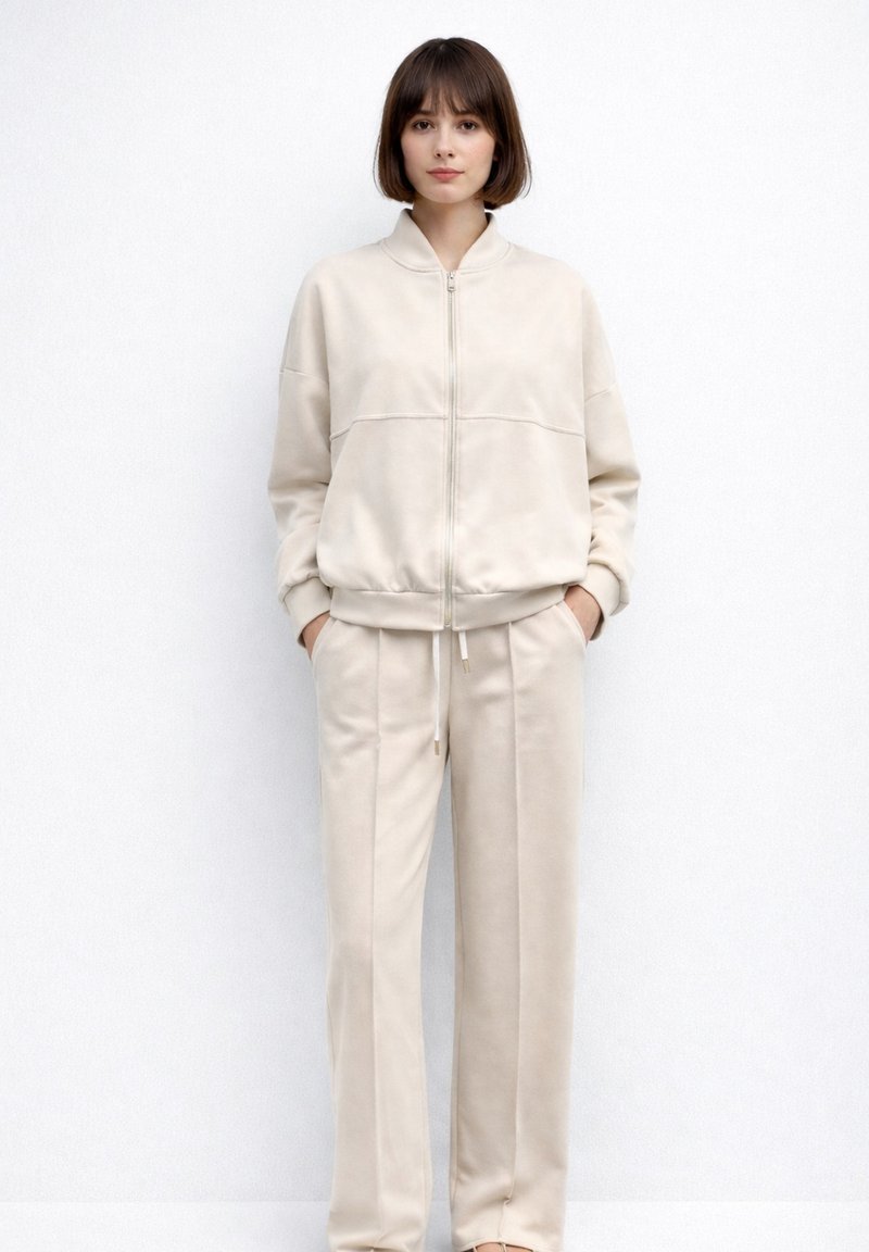 Ung kvinde stående med hænderne i lommerne iført beige lynlåstrøje og matchende bukser med vide ben foran en ensfarvet hvid baggrund.