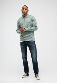 Polo in Mintgrün mit langen Ärmeln, Kragen, drei Knöpfen und einem Logo. Kombiniert mit dunkelblauen Jeans und schwarzen High-Top-Sneakern.