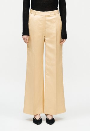 Személy magas derekú, bőszárú, bézs nadrágot visel fekete hosszú ujjú felsővel és fekete hegyes orrú cipővel.