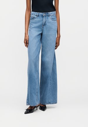 Nő világoskék, magasderekú, bőszárú farmert visel, fekete, hegyesorrú kis sarkú cipővel és fekete ujjatlan felsővel párosítva.