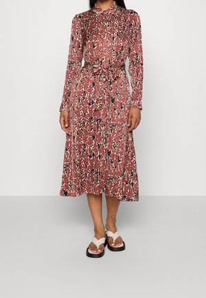 Kvinde iført en langærmet midi-kjole med rødt, sort og beige abstrakt mønster, bundet taljebælte og hvide plateau-sandaler.