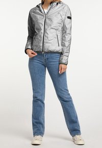 Zilveren, lichtgewicht jas met capuchon en een getextureerd oppervlak. Voorzien van ritszakken en een aansluitende zoom. Gecombineerd met blauwe spijkerbroek en witte sneakers.