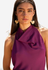 Top morado sin mangas con un escote drapeado, hecho de una tela suave. La modelo lleva pendientes dorados con flores. Fondo simple, enfatizando el atuendo.