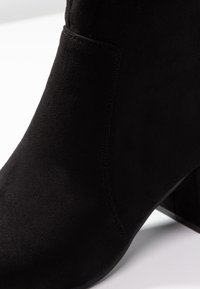 Primo piano di un stivale alla caviglia in suede nero con tacco a blocco e cuciture visibili su una superficie bianca.