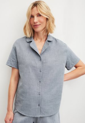 Femme blonde portant une chemise à manches courtes bleu clair avec un col, debout devant un fond blanc.
